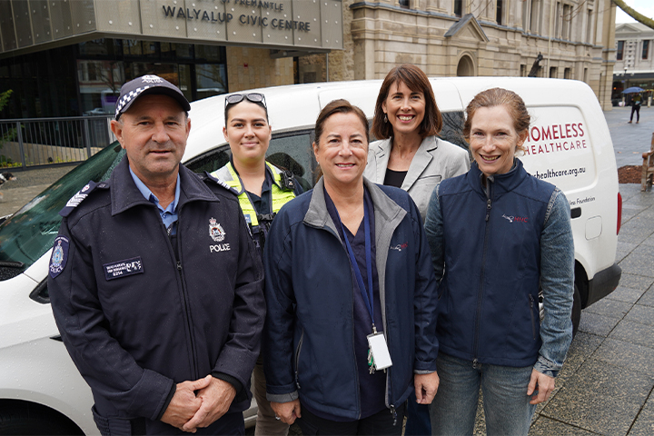 Five people, including two police officers and three women in jackets, stand smiling in front of a Homeless Healthcare van parked outside the Fremantle Walyalup Civic Centre.