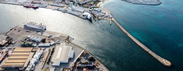 Aerial view of Fremantle harbor with warehouses, shipping containers, and boats docked along the shoreline. A long pier extends into clear blue-green water, with a marina and city buildings visible in the background.