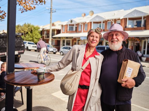 An older woman in a headscarf and red outfit stands smiling beside an older man in a pale hat, holding a bag and a coffee, on a sunny street with houses, cars, and outdoor tables.