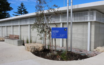 Modern concrete building with decorative white metal panels. In front, a blue sign reads South Beach Changerooms near two flagpoles, benches, rocks, and small landscaped plants. Trees and a blue sky are in the background.