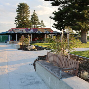 A modern outdoor café with colorful signage is surrounded by tall pine trees and greenery. In the foreground, a wooden bench sits beside a concrete walkway under the soft sunlight.