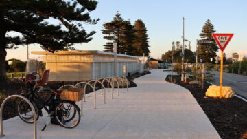 Sunlit pathway with bike racks, a parked bicycle with a basket, tall pine trees, a modern building, and a Give Way traffic sign near a road. The area appears clean and well-maintained.