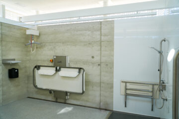 A modern, spacious accessible bathroom with a fold-down adult changing table, wall-mounted shower, grab bars, and a bench. The room has light-colored walls and a non-slip floor.