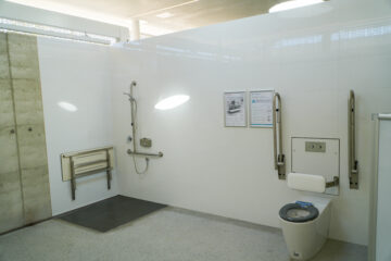 Accessible public restroom with a toilet, fold-up support rails, wall-mounted backrest, roll-in shower with a fold-down seat, handheld showerhead, and informational posters on the white tiled wall.
