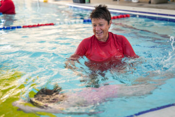 A swim instructor in a red shirt smiles while guiding a young child who is swimming underwater in an indoor pool. Blue lane dividers and poolside can be seen in the background.