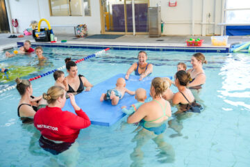 A group of adults and babies participate in a swim class in an indoor pool. An instructor in a red shirt leads the session as babies lie on a blue floating mat surrounded by smiling adults in the water.