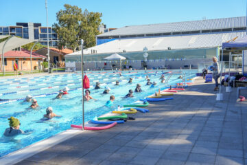 A group of adults participate in a water aerobics class in an outdoor pool on a sunny day, with colorful pool noodles lined up along the pool deck. An instructor stands poolside giving directions.