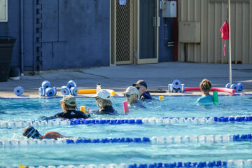 Adults participate in a water aerobics class in an outdoor pool, using colorful foam noodles and wearing hats. Pool equipment is visible on the deck, and lane dividers mark sections of the pool.