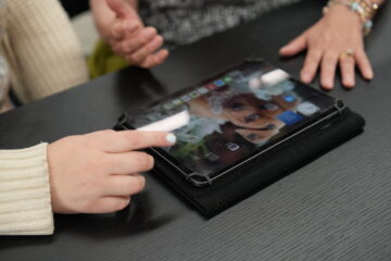 Two people sit at a table with a tablet in front of them. One person points at the tablet’s screen, which displays a wallpaper image of a girl and app icons. Both have their hands visible near the device.
