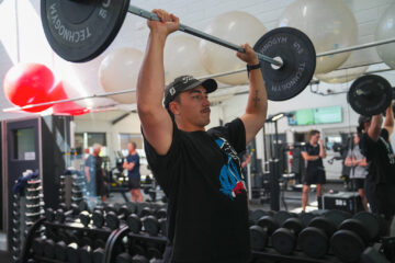 A person wearing a black T-shirt and cap lifts a barbell overhead in a gym, with others exercising in the background and large exercise balls on a wall shelf.