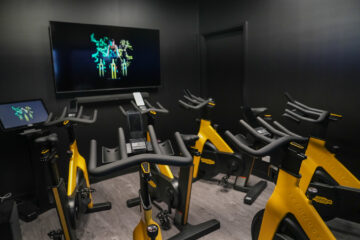 A small, dark indoor cycling studio with several yellow exercise bikes facing a large screen displaying colorful graphics.