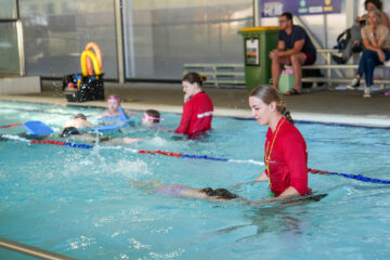 A swimming instructor in a red shirt helps a child in a pool while other children practice nearby. Adults sit on benches in the background, watching the lesson. Pool equipment and a trash bin are visible.