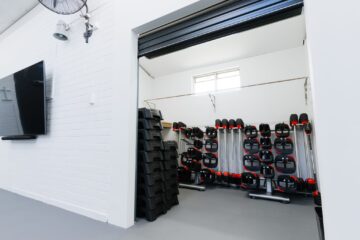 A fitness studio storage room with stacked aerobic steps and shelves holding barbells with red weights. A wall fan and a TV are mounted on the white brick wall outside the open doorway.