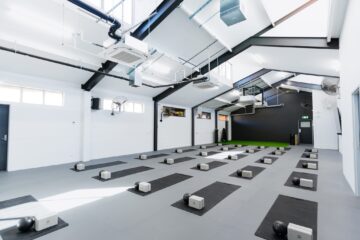 A bright, spacious fitness studio with black yoga mats and neatly arranged yoga blocks on the floor, under a high ceiling with exposed beams and skylights.