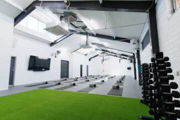 A modern gym with yoga mats and blocks neatly arranged on the floor, a green turf area, a large TV on the wall, and dumbbells stacked on racks. The room has white walls and high ceilings.
