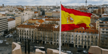 The Spanish flag waves prominently in the foreground with a cityscape of Madrid, featuring historic buildings and dense rooftops, under a cloudy sky in the background.
