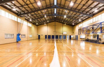 An empty indoor gymnasium with a wooden floor, high ceiling, visible court markings, a volleyball net in the center, basketball hoops, and rows of chairs along the right wall.