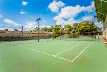 An empty outdoor tennis court with green surface, surrounded by trees, a building, and a fence, under a partly cloudy blue sky during the day.