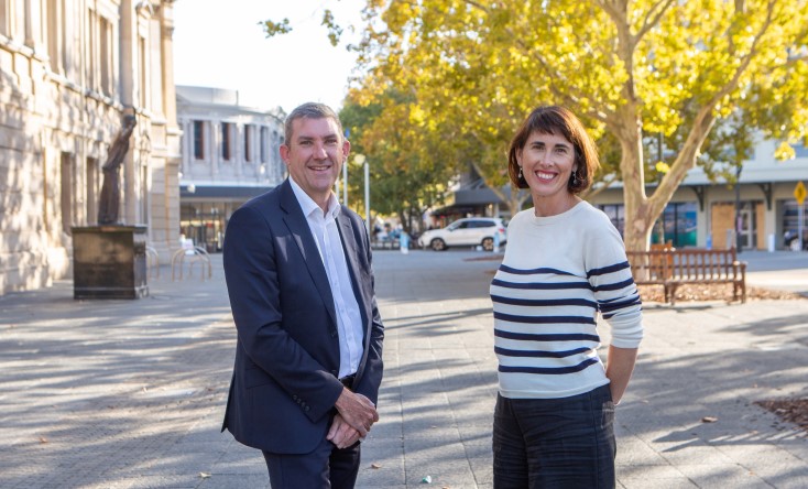 A man in a suit and a woman in a striped sweater stand smiling on a sunny city street with trees and buildings in the background.