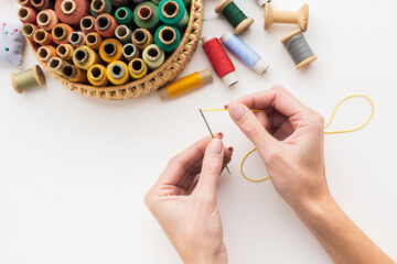 Two hands thread a needle with yellow thread next to a basket filled with colorful spools of thread and several loose spools on a white surface.