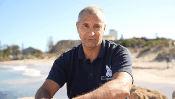 A man wearing a navy blue City of Fremantle polo shirt sits outdoors on a sunny beach, with sand, water, and distant trees and buildings in the background.