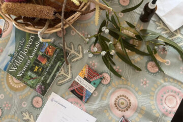 A table with a basket of native plants, a Guide to the Wildflowers of Western Australia book, a branch with leaves, an Aboriginal artwork card, a spray bottle, a notepad, and patterned tablecloth.