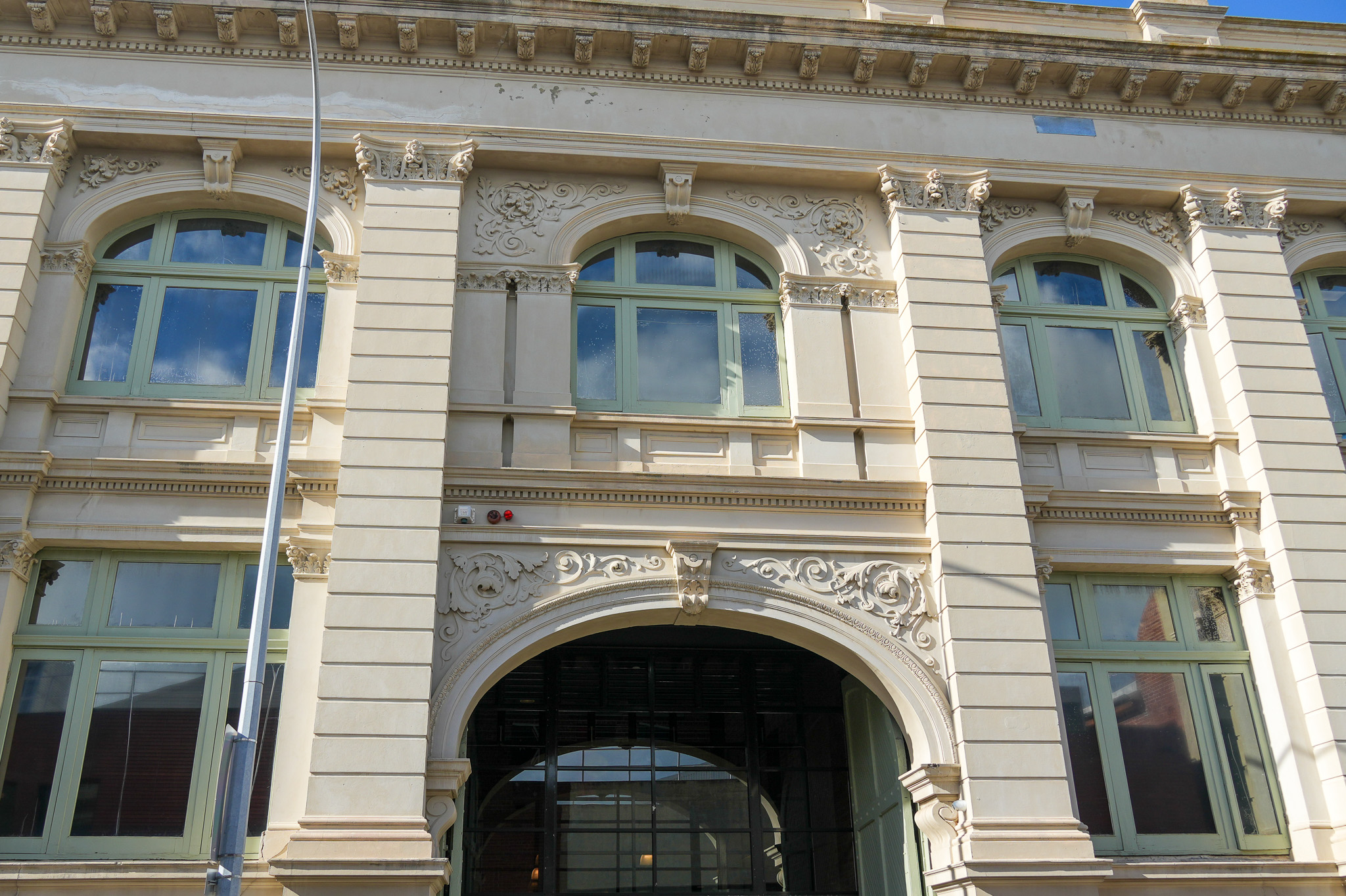 Ornate beige building facade with large arched windows, decorative carvings above an arched entrance, and reflections of blue sky and clouds in the glass.