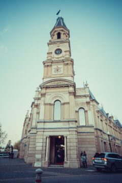 A tall, beige clock tower with arched windows stands on a street corner. People are entering the building, and parked cars are nearby. The sky above is clear with a soft blue tint.