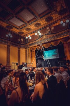 A large group of people in formal attire are seated at round tables in an ornate hall with high ceilings and decorative moldings. A performer on an aerial hoop is suspended above the stage near a screen displaying event details.