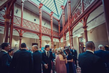 A group of people dressed in formal attire gather and converse in a large, elegant hall with a glass ceiling and red wooden railings.