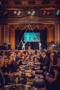 A formal banquet hall filled with seated guests watching two acrobats perform on a tall metal structure on stage, with a “Fremantle Business Awards” sign displayed in the background.