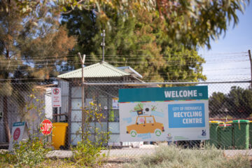 A sign reading Welcome to City of Fremantle Recycling Centre is attached to a chain-link fence with bins and a small building visible behind, surrounded by trees and greenery.