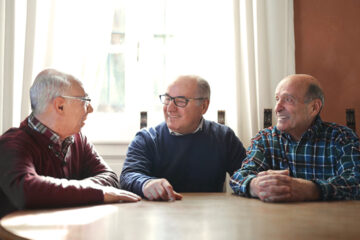 Three elderly men sit together at a wooden table, smiling and talking, with sunlight streaming in through a window behind them.