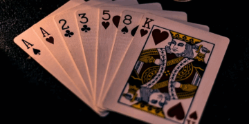 A close-up of seven playing cards fanned out on a dark surface: Ace of spades, Ace of clubs, 2 of clubs, 3 of spades, 5 of hearts, 8 of hearts, and King of hearts.