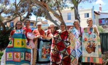Seven women stand outdoors, smiling and holding colorful handmade quilts with various patterns, including numbers, geometric designs, and a teddy bear in a basket with balloons. Trees and buildings are visible in the background.