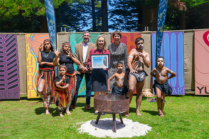 A group of adults and children, some in traditional Indigenous attire, stand together outdoors on grass in front of colorful banners. A woman in the center holds a framed certificate. A fire pit is in the foreground.