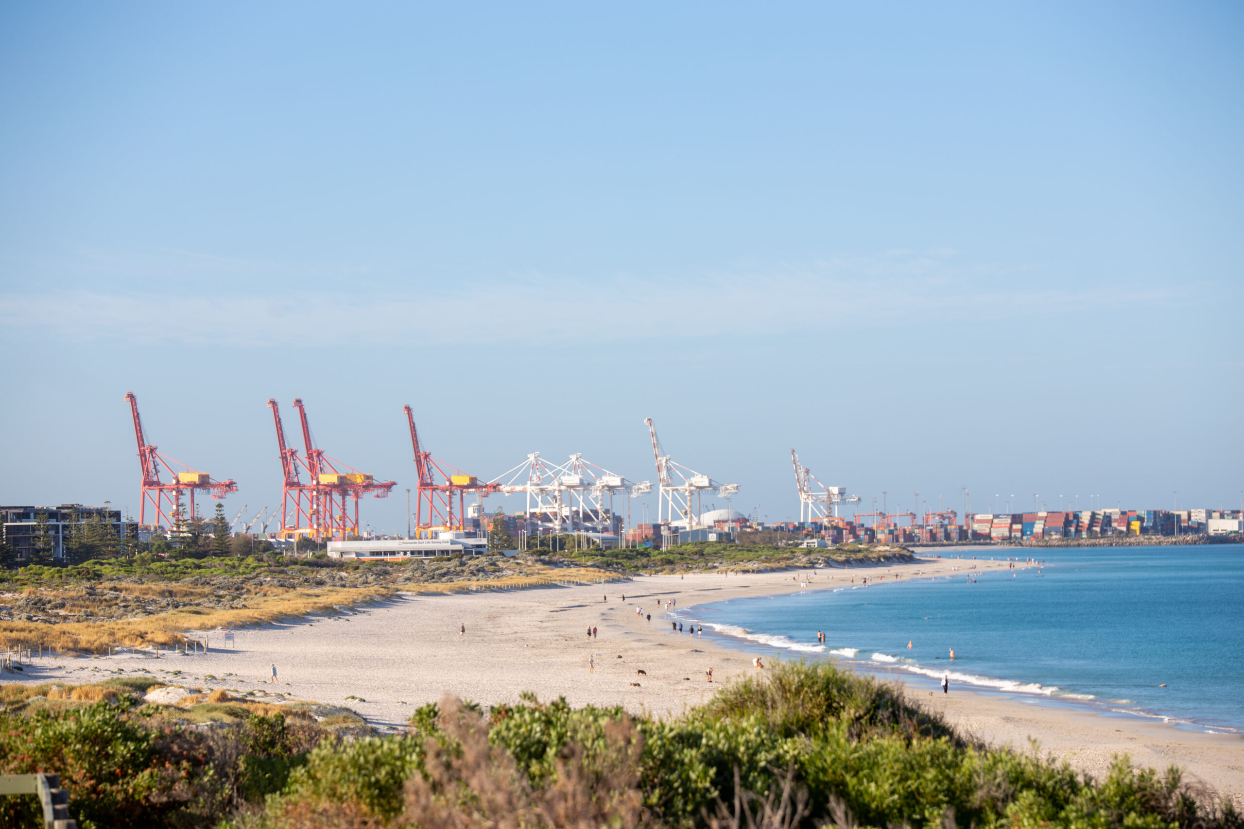 A sandy beach with scattered people walking along the shore. Beyond the beach, large red and white shipping cranes stand at a busy port, with containers and blue sky in the background.