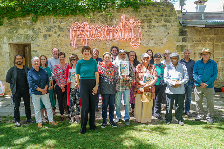 A group of people stand and smile outdoors in front of a stone wall with a neon “Moorditj” sign. Some hold certificates or books, and trees provide shade overhead.