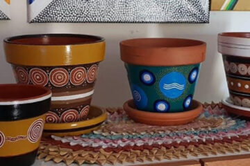 Four terracotta pots decorated with colorful Aboriginal dot painting designs are displayed on a shelf with woven mats and other artwork in the background.