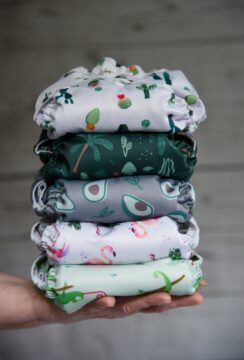 A hand holds a stack of six colorful cloth diapers, each featuring different patterns such as plants, avocados, trees, and whimsical designs, against a wooden background.