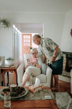 An elderly woman sits in an armchair looking at her phone, while an elderly man leans over her shoulder to see the screen. They are in a cozy, well-lit living room with wooden furniture and houseplants.