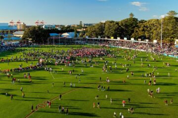 A large crowd of people, many dressed in red, gather and walk across a green sports field under a clear blue sky, with buildings, trees, and stadium lights in the background.