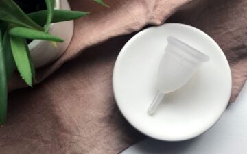 A white menstrual cup rests on a round white plate, placed on a light brown cloth next to a green potted succulent plant.