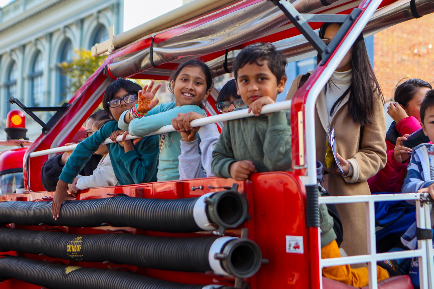 A group of smiling children ride on the back of a red fire truck, holding onto the railing and waving, while adults stand behind them. The scene appears festive and joyful on a city street.