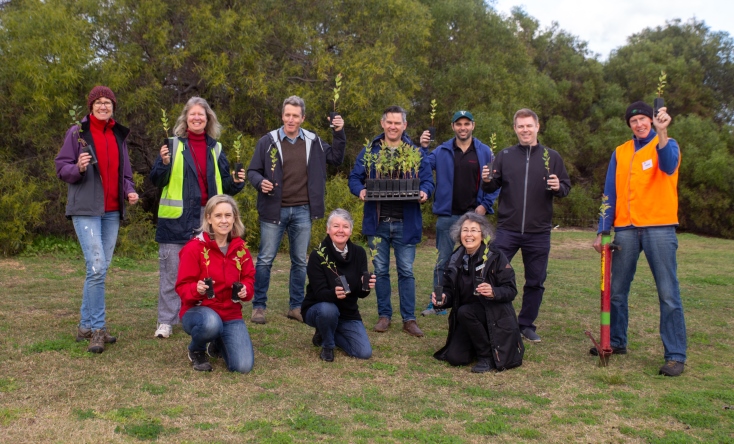 A group of eleven people outdoors, some kneeling and some standing, smiling and holding small tree saplings, with green bushes and trees in the background.