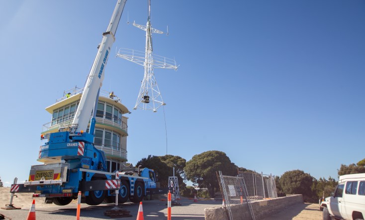 A crane lifts a large metal radio tower next to a multi-story building under clear blue skies. Orange safety cones and fencing mark the construction area, with trees and vehicles nearby.