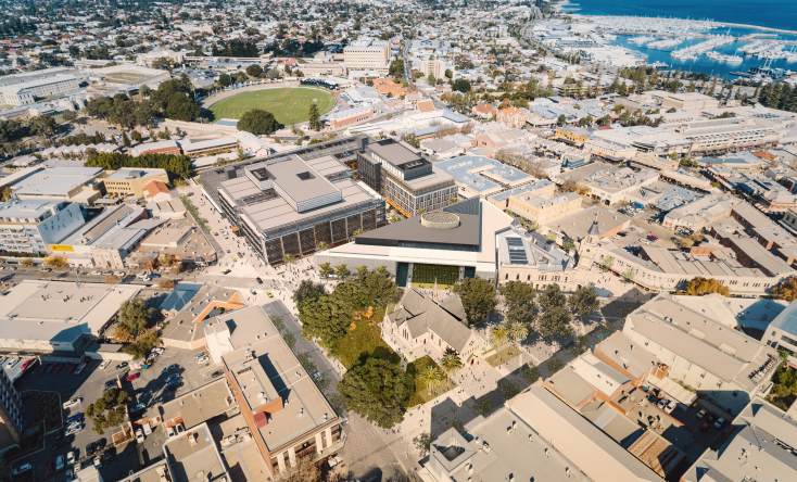 Aerial view of a coastal city with a mix of modern and historic buildings, a sports field, tree-lined streets, and a marina with boats by the waterfront.