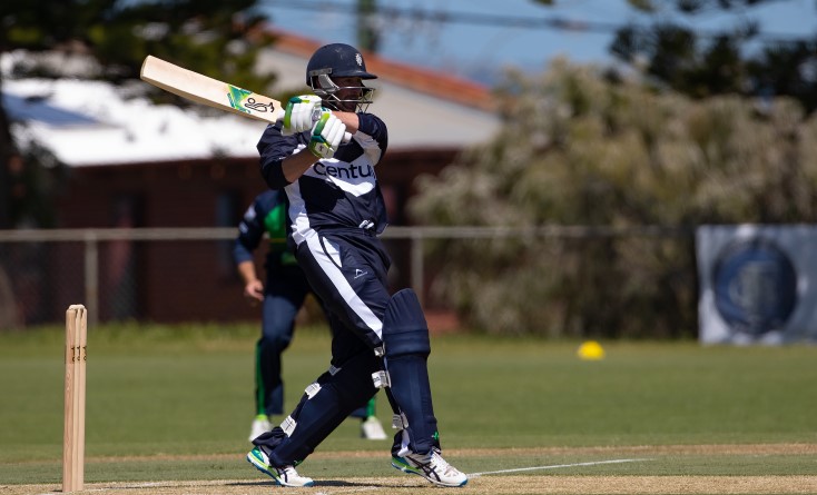 A cricket player in navy blue gear swings his bat while batting during a match on a sunny outdoor field. Another player in dark uniform stands behind him. Trees and a building are in the background.