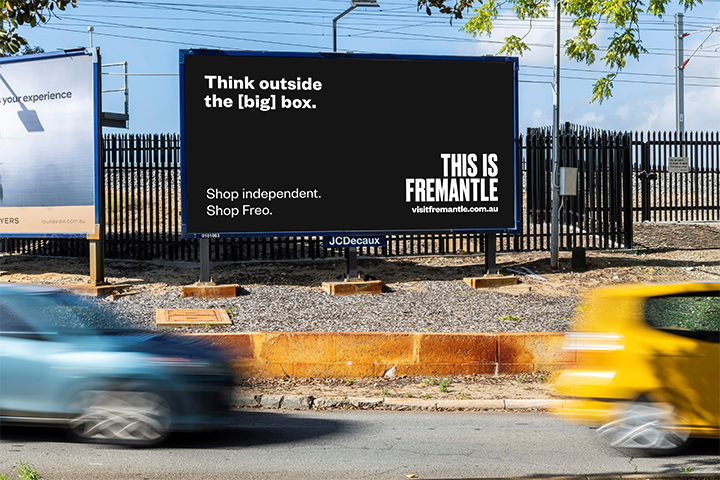 A black billboard beside a road reads, Think outside the [big] box. Shop independent. Shop Freo. THIS IS FREMANTLE visitfremantle.com.au. Blurred cars drive past in the foreground.