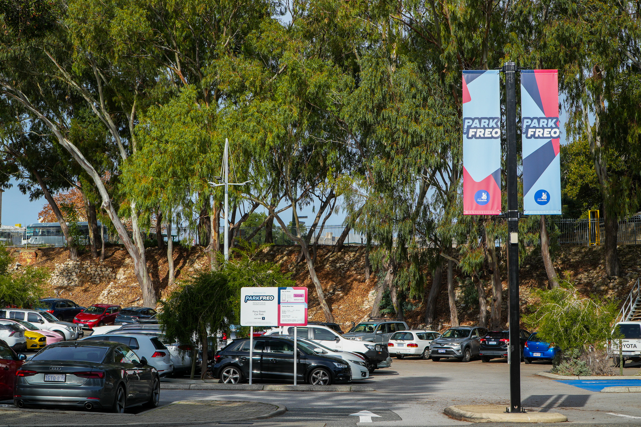 A parking lot filled with cars is bordered by tall trees. Two vertical banners on a pole display “PARK FREO.” Signs and vehicles are visible throughout the lot under a clear, sunny sky.